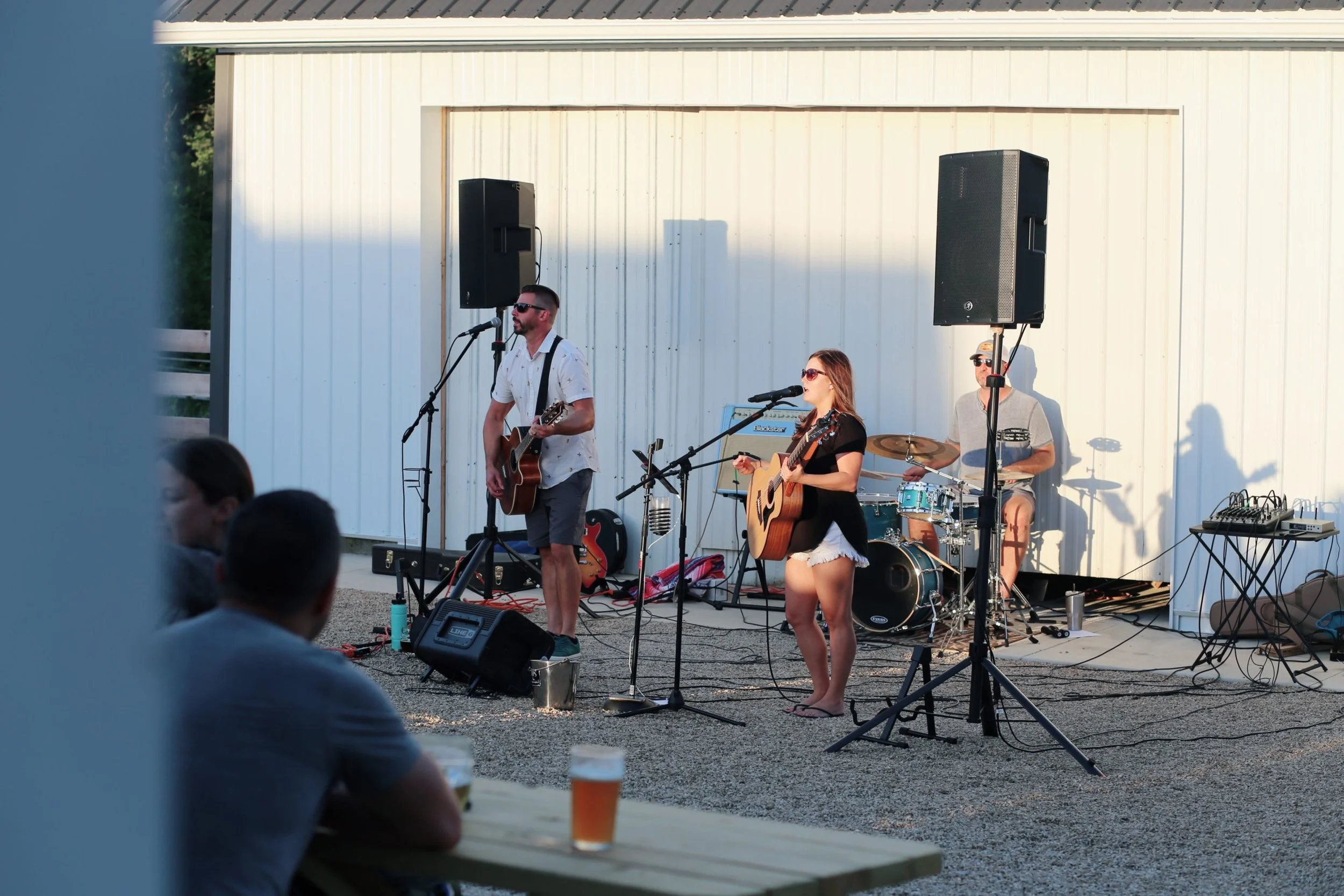 A musical trio performs on an outdoor gravel stage in front of a white building.