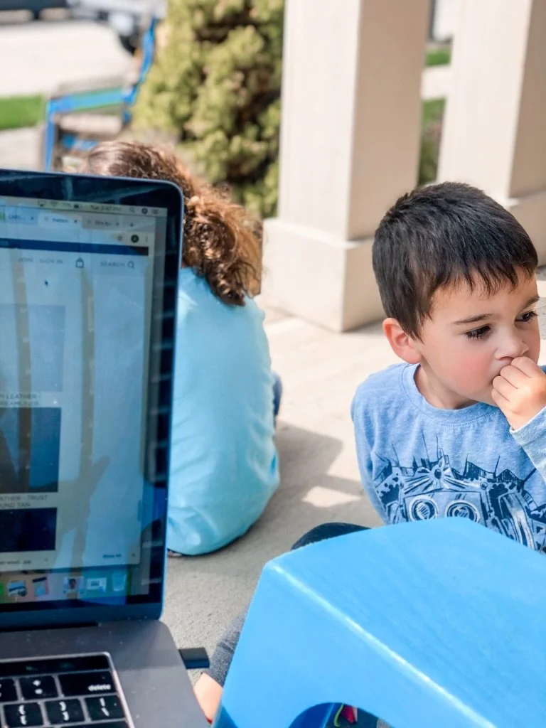 A boy sits near a laptop screen showing a retail website while another child sits in the background.