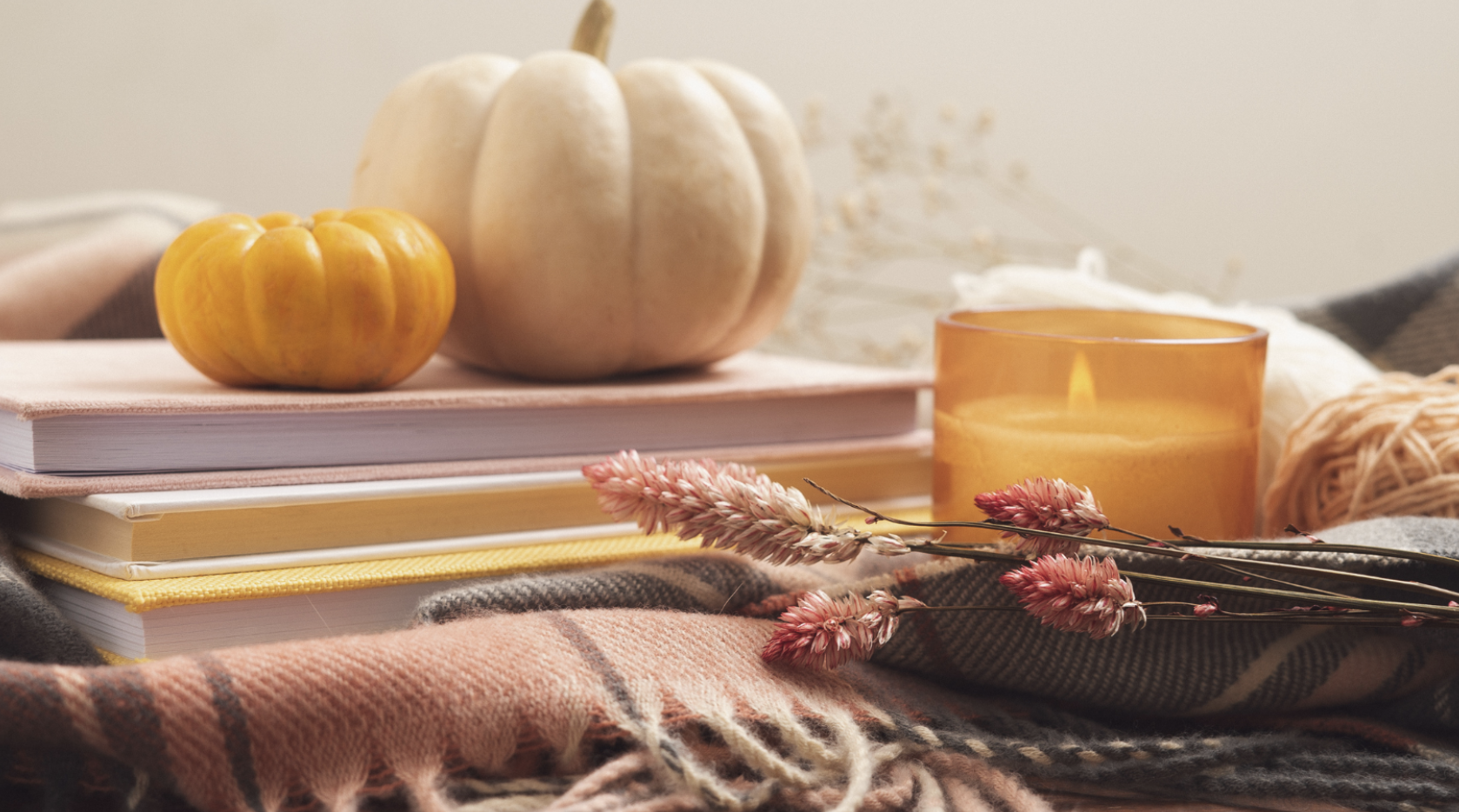 A stack of books topped with two pumpkins, a lit candle, and dried flowers on a plaid blanket.