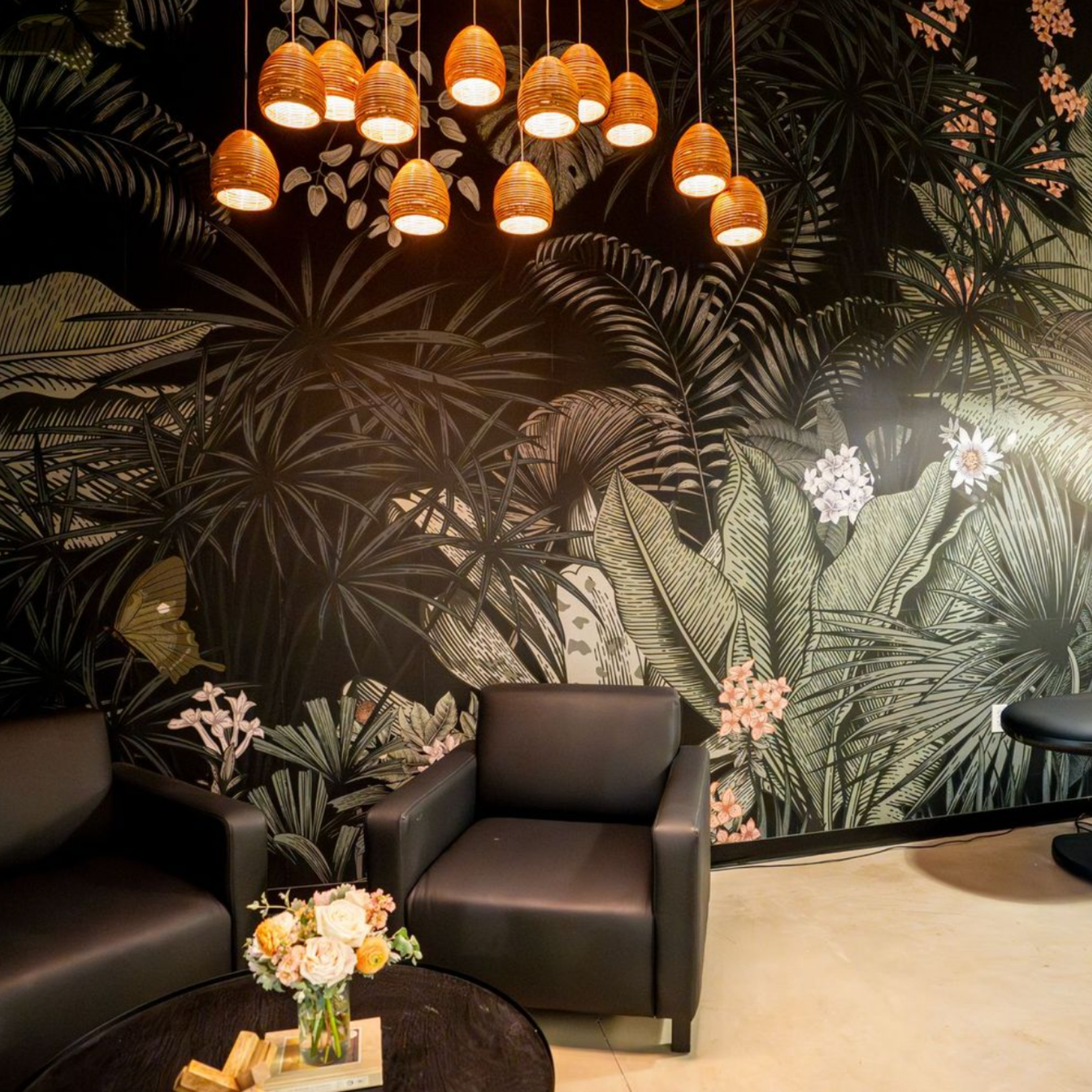 Two armchairs facing a round coffee table in front of a wall with dark tropical-themed wallpaper and pendant lights.