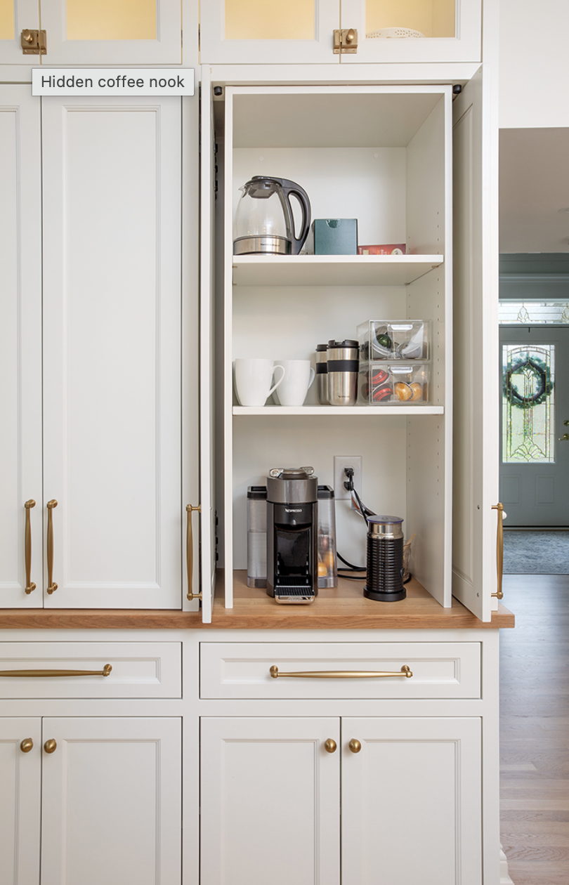 Open cabinet used as a coffee station with an electric kettle, coffee machine, milk frother, and mugs on shelves.