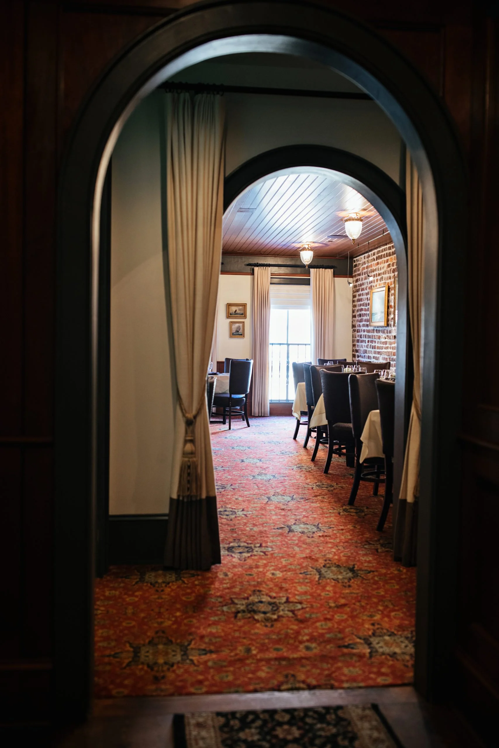A view through an arched doorway into a dining room with tables, chairs, and patterned red carpet.