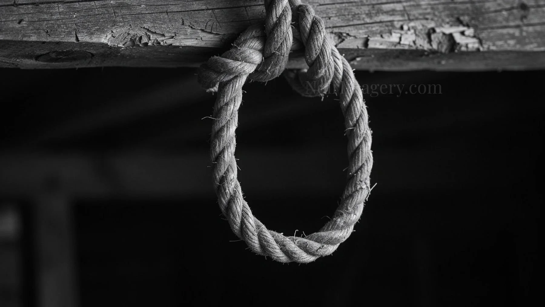 Black and white image of a rope noose hanging from a wooden beam in a dark environment