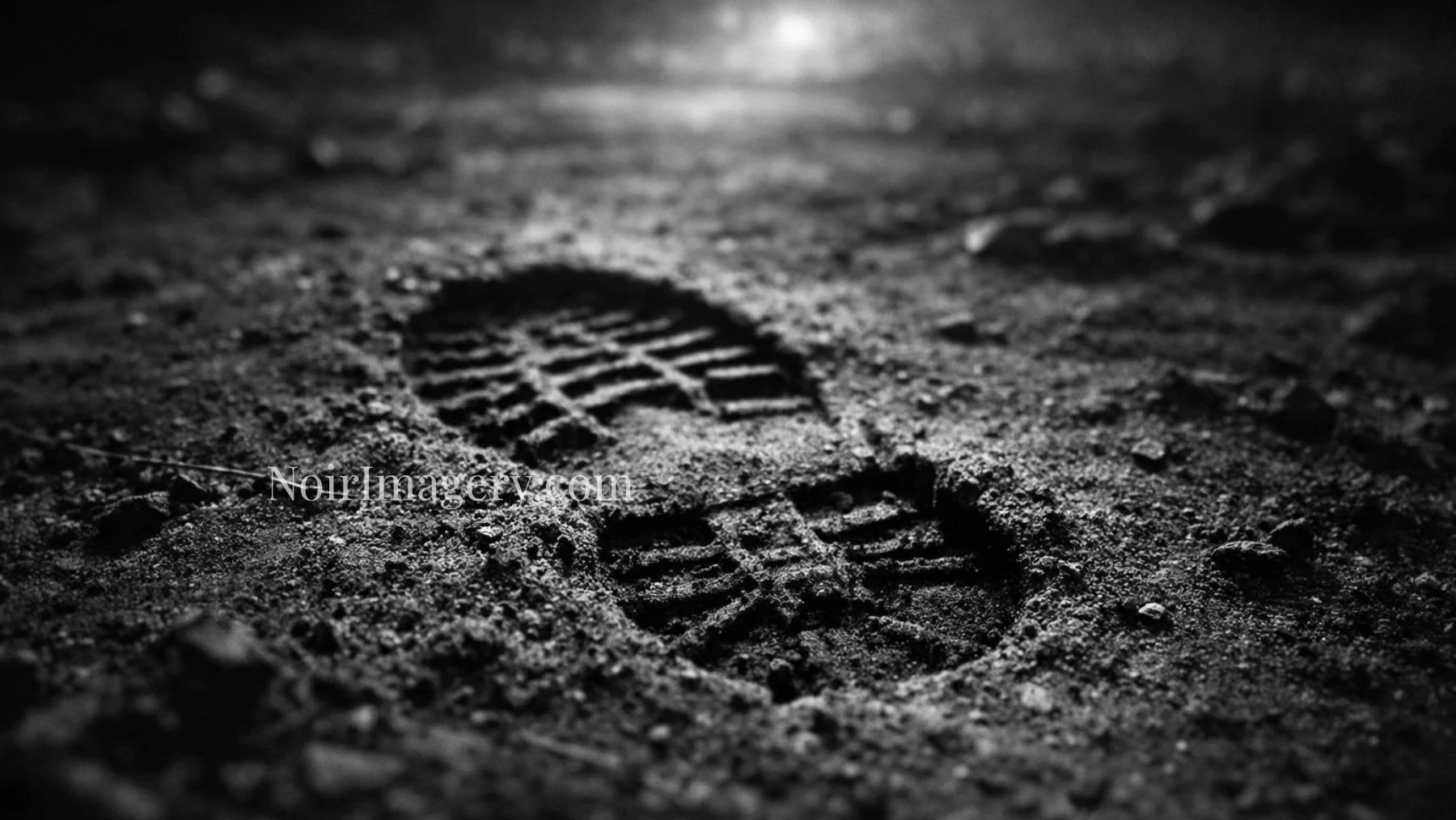 Black and white noir image of a boot footprint pressed into dirt with dramatic lighting.