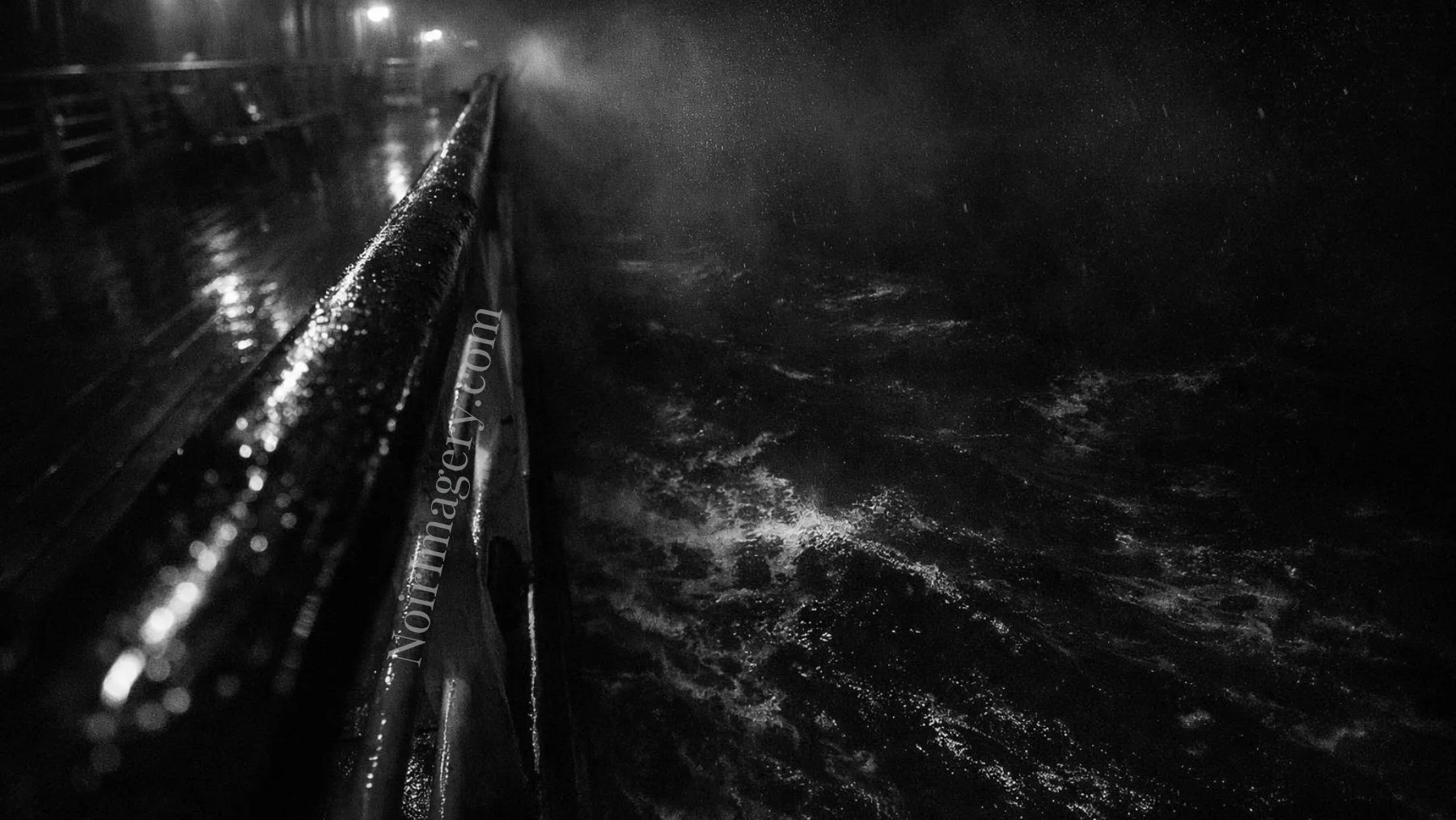 Black-and-white image of rough ocean water beside a rain-covered ship railing at night.