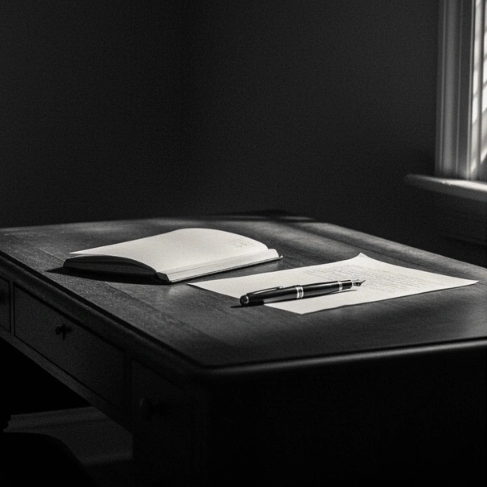 Small writing desk with papers and pen near window with venetian blinds.