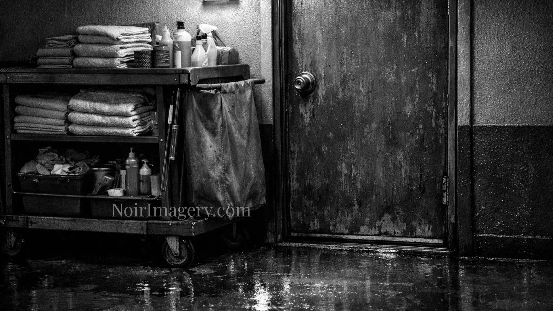 Black-and-white image of a motel housekeeping cart parked beside a weathered guest room door on a wet walkway.
