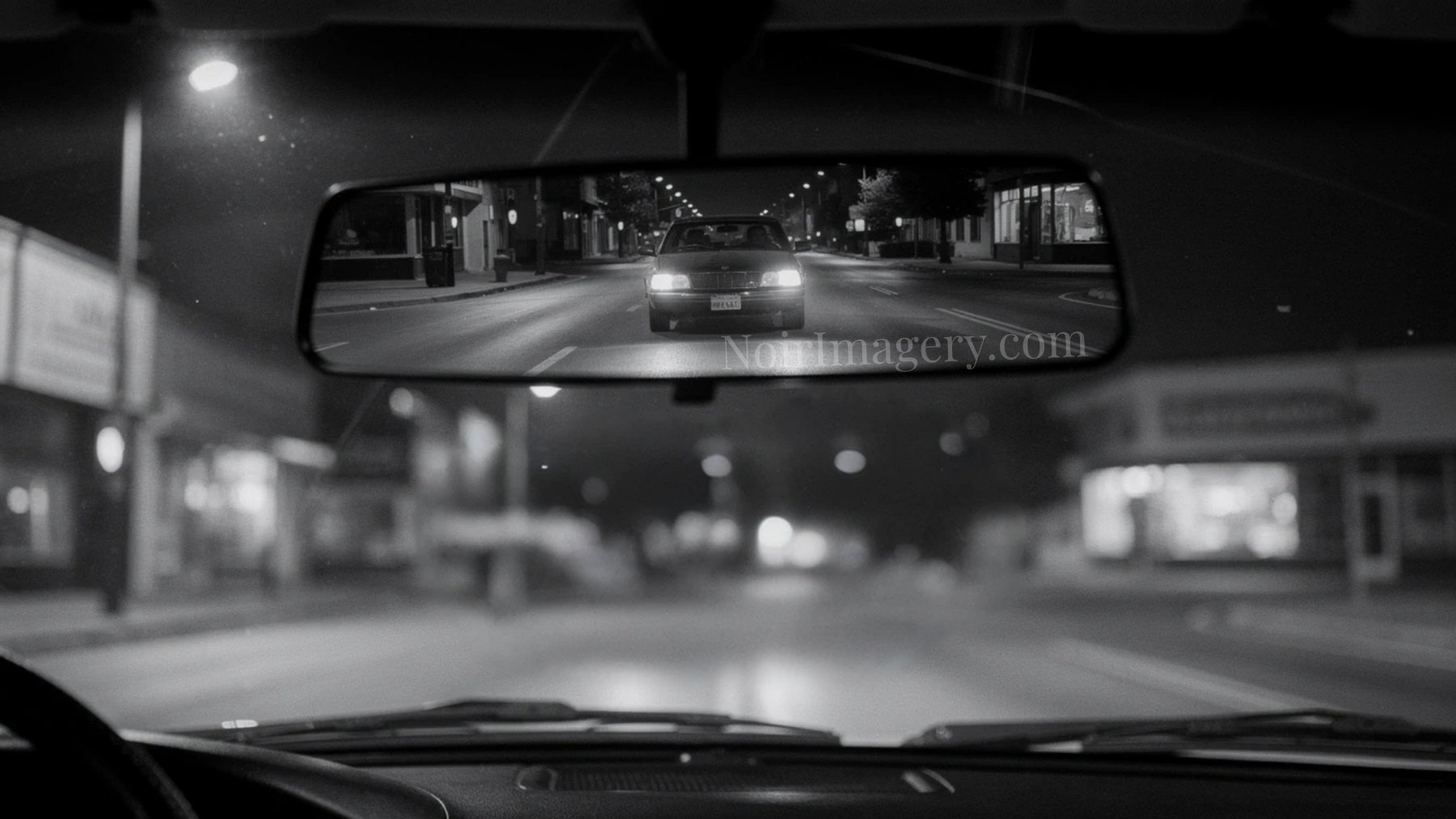 Black-and-white noir image of a trailing car reflected in a rearview mirror at night.