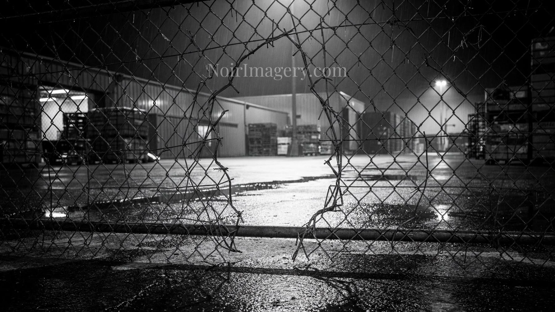 Black-and-white image of a chain-link fence with a large section cut away, creating an opening into an industrial yard.
