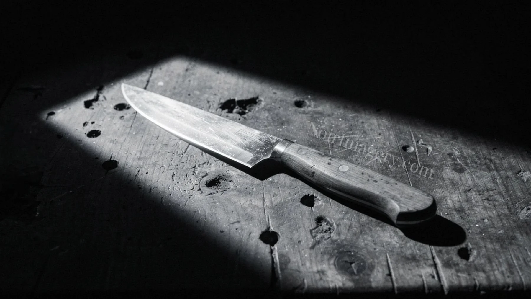 Black and white cinematic image of a sharp knife lying on a worn wooden surface in dramatic shadow