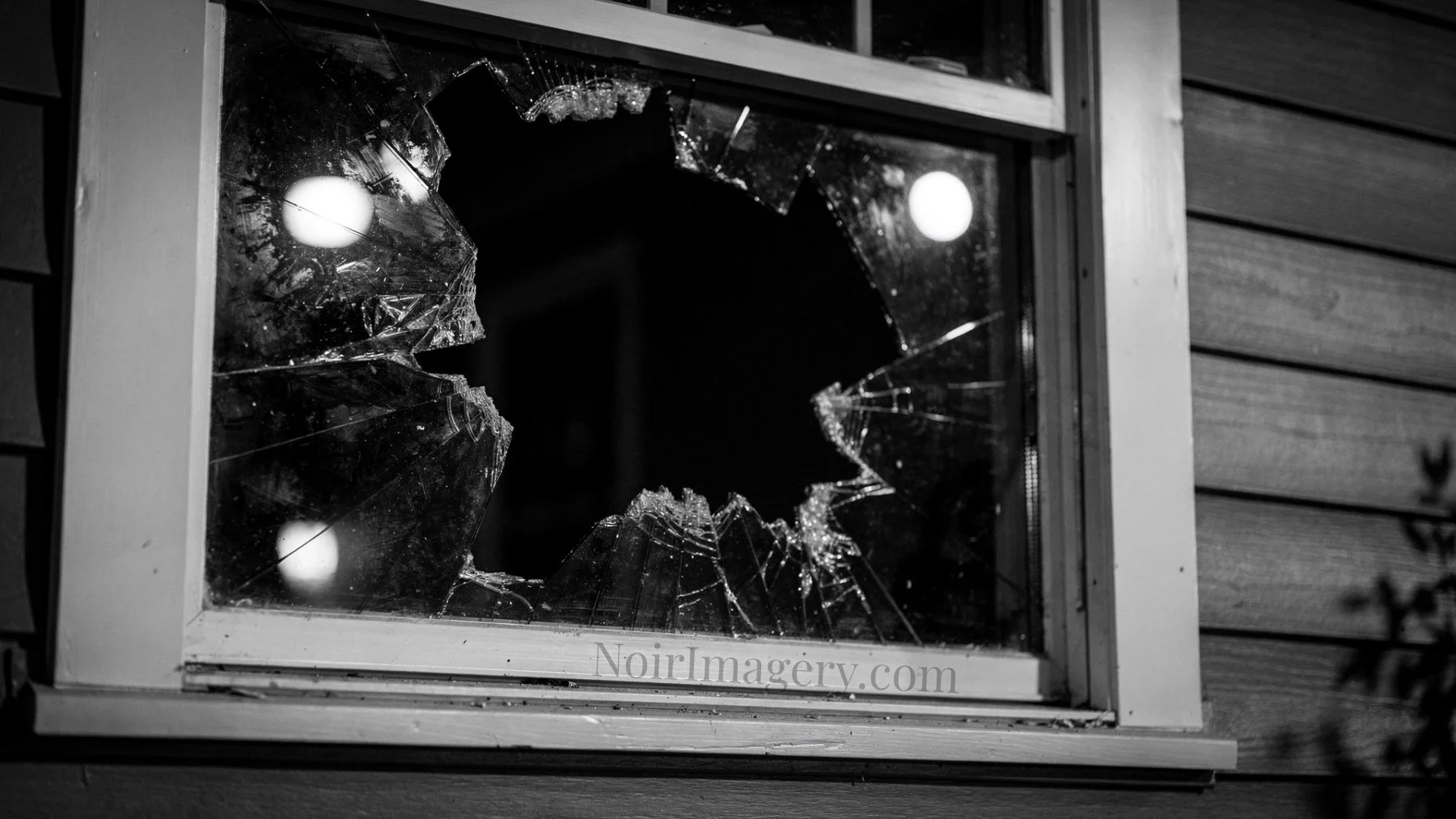 Black-and-white image of a broken house window with shattered glass and a large opening in the pane at night.