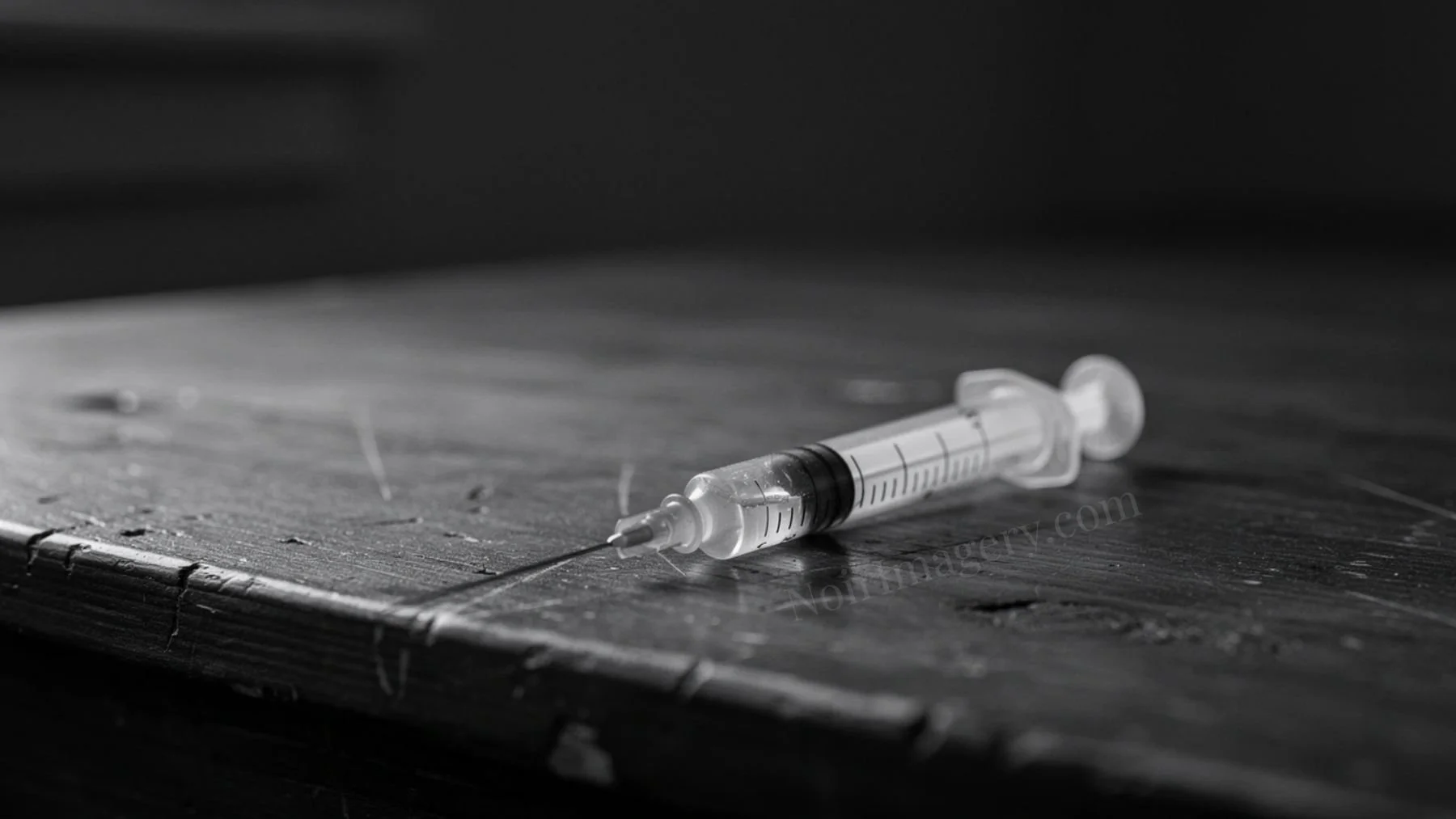 Black and white cinematic image of a syringe lying on a rough wooden table with dramatic lighting