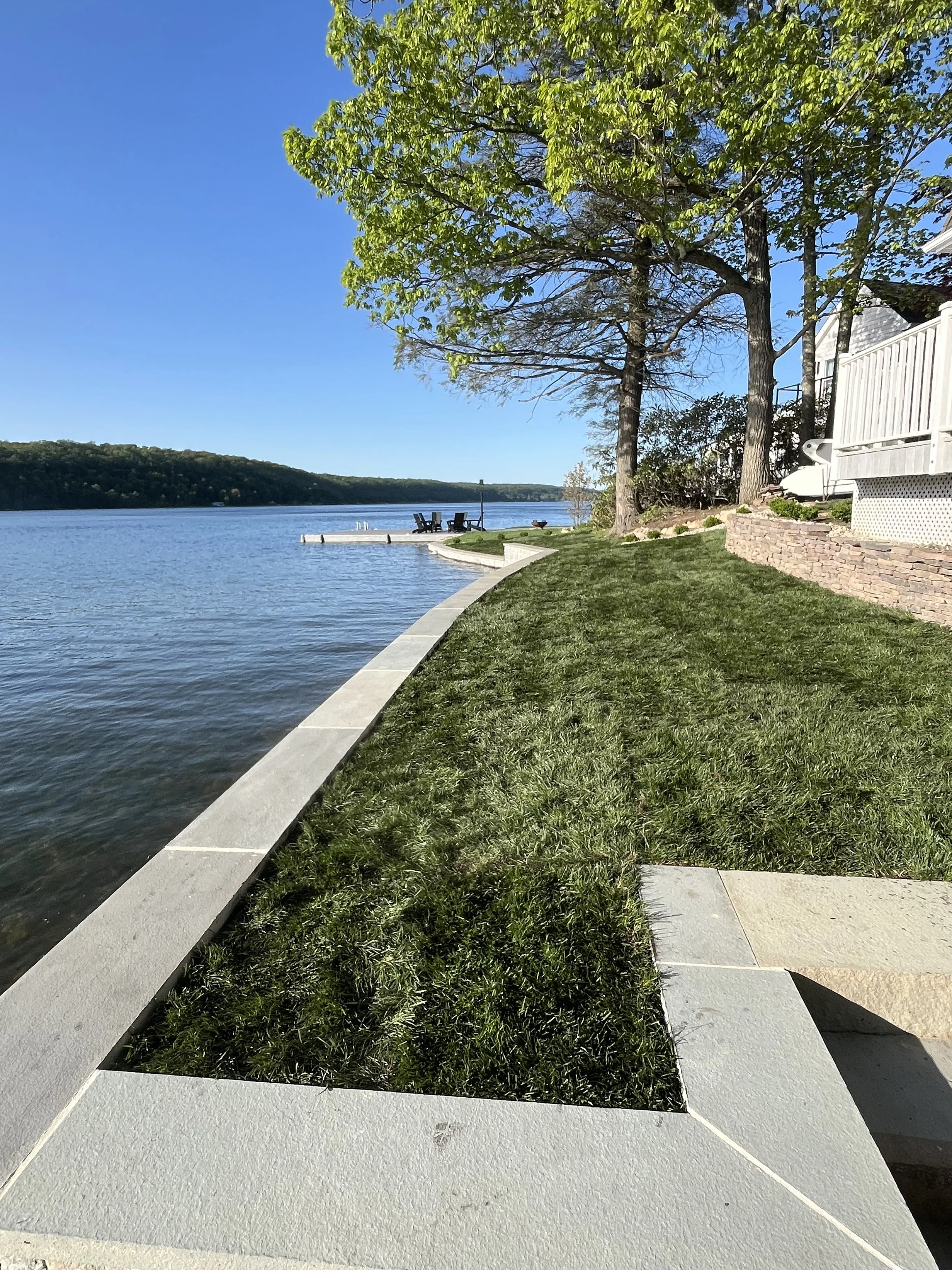 View of a lakeside yard with a stone border, trimmed grass, and trees. A dock with chairs extends into the water, with a calm lake and distant hills in the background.