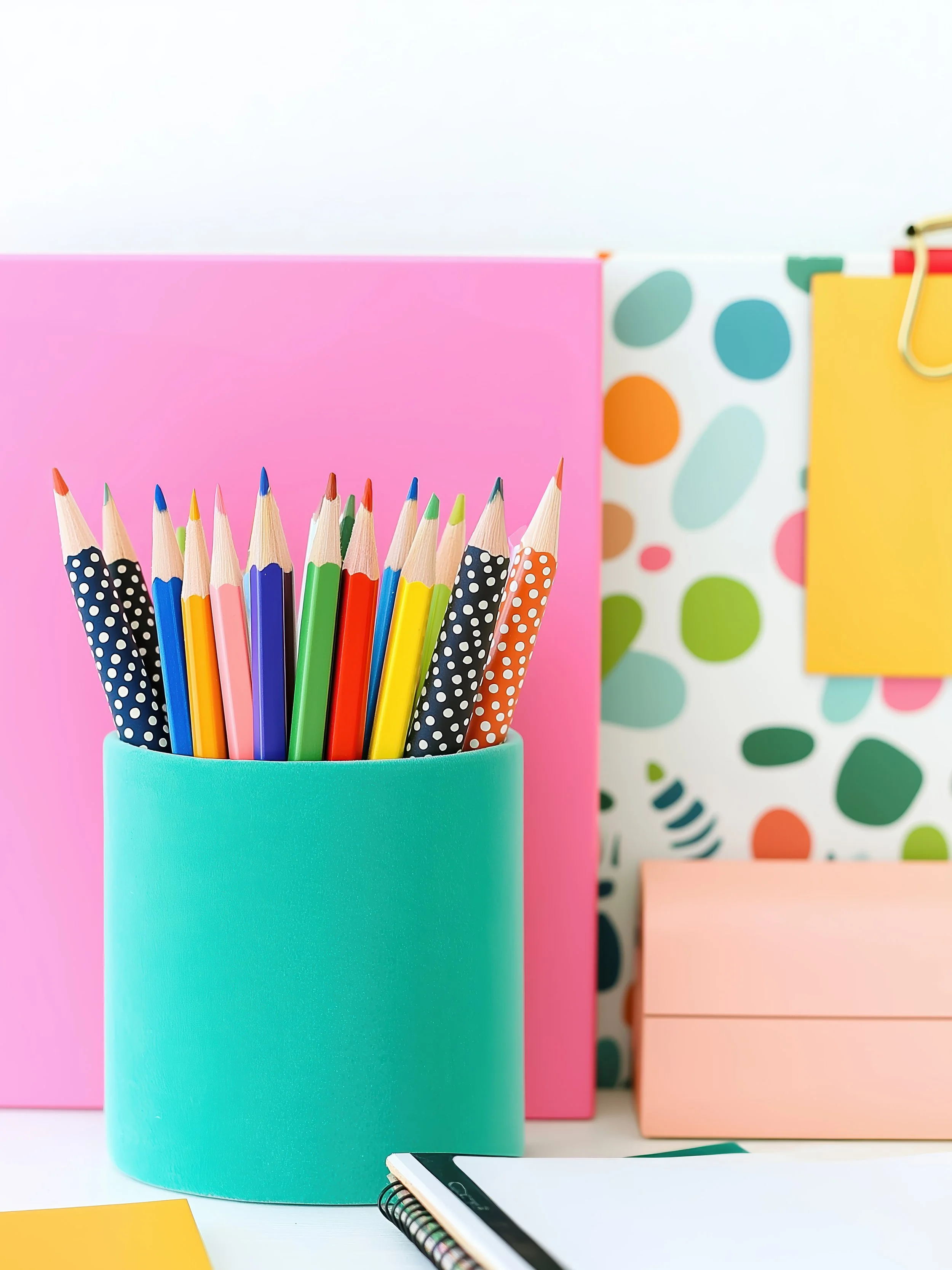 Colorful pencils in a teal holder on a desk with pink, yellow, and patterned notebooks and a spiral-bound notebook in the background.