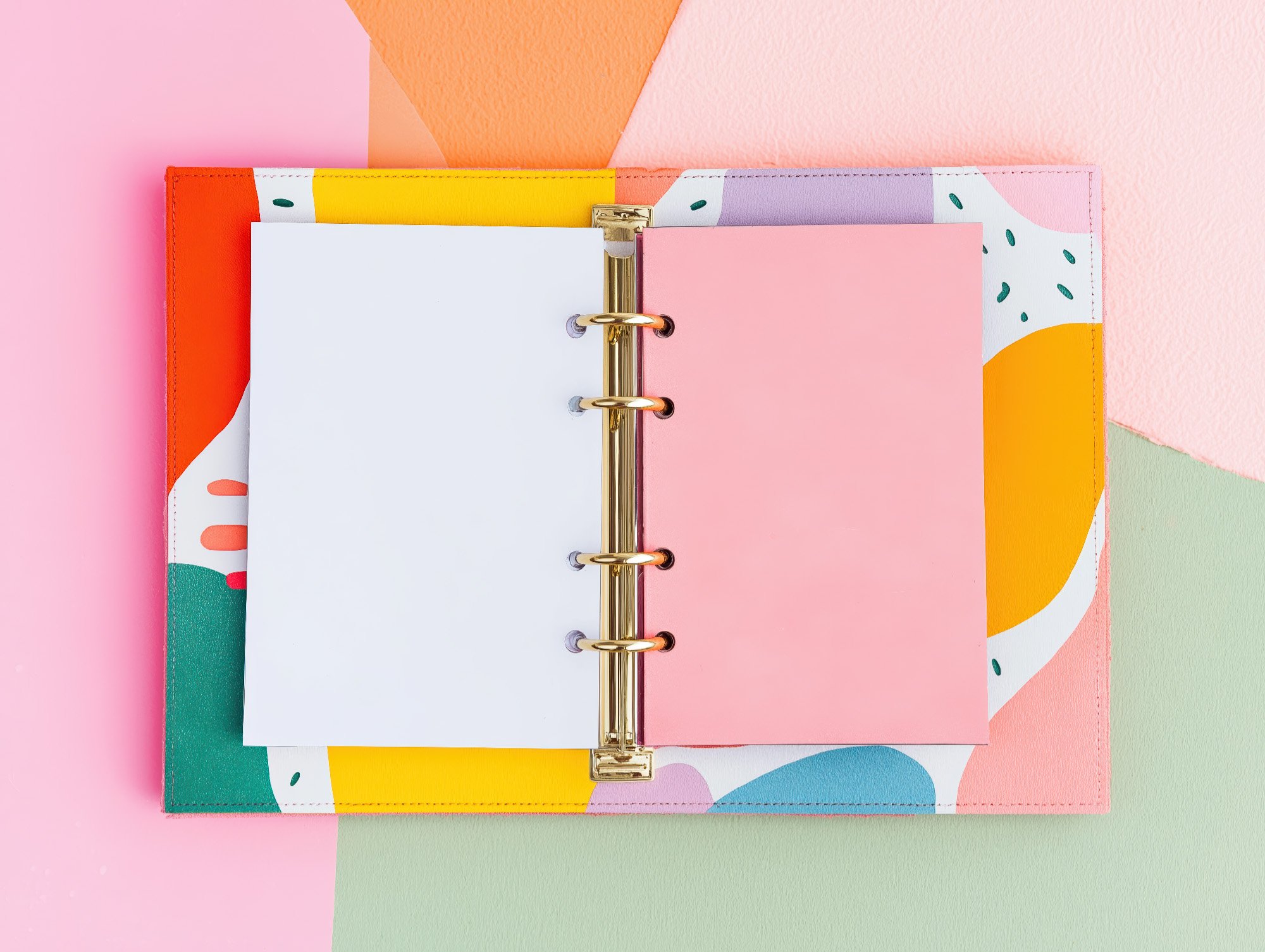 Open colorful planner with blank white and pink pages, gold binder rings, on pastel pink, green, peach, and light purple backgrounds.