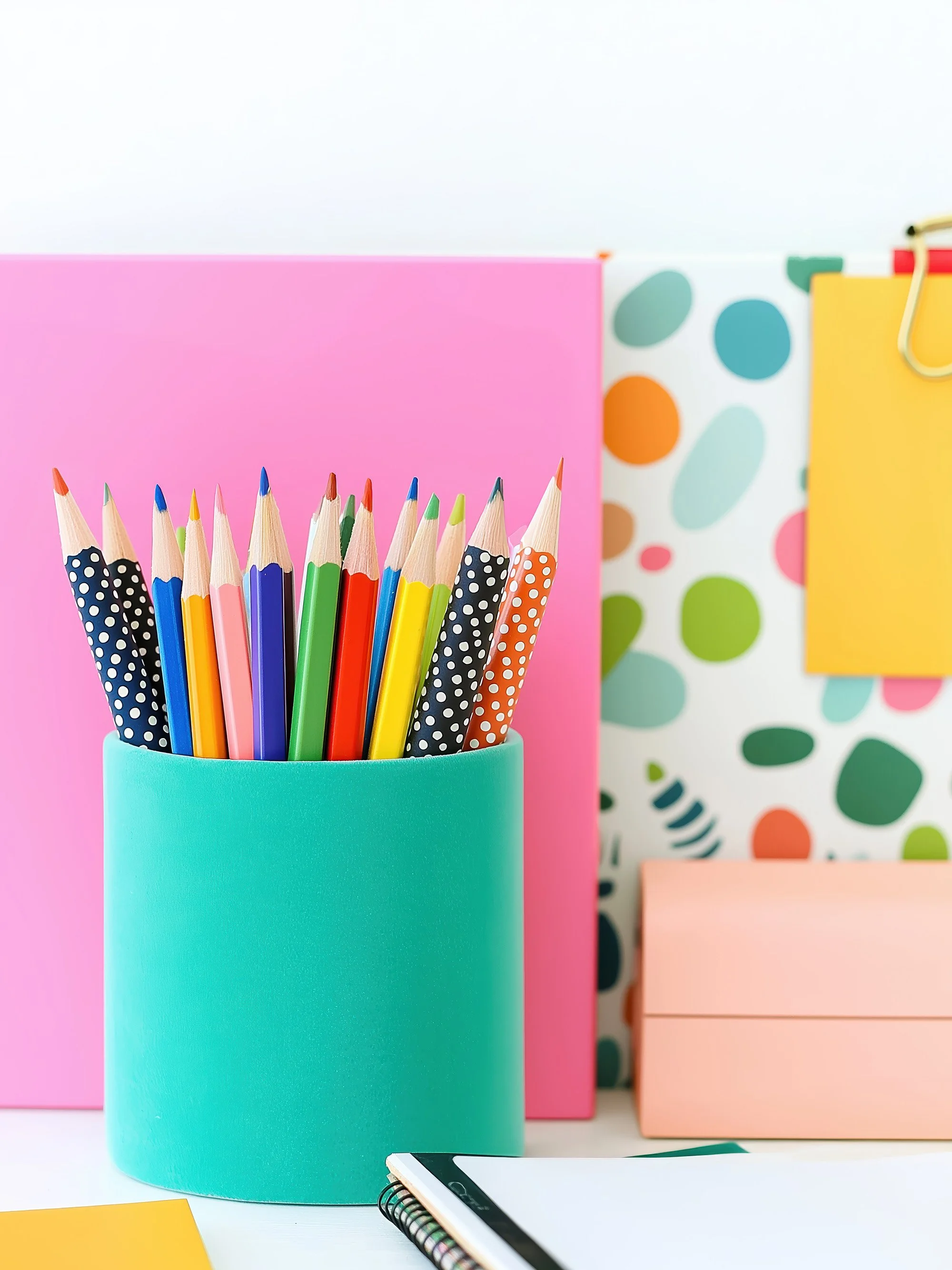 A colorful collection of pencils in a teal holder, with pastel-colored boxes and a patterned background of dots and shapes.