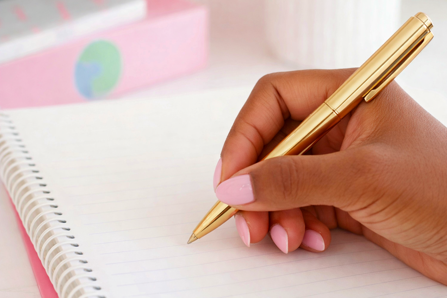A person with pink manicured nails holding a gold pen and writing in a spiral notebook with lined paper.