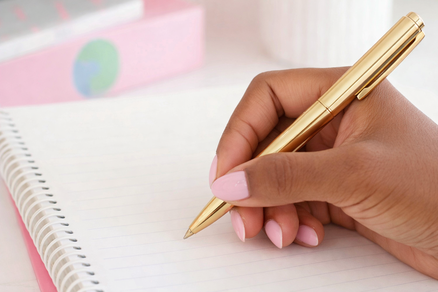 A person with pink nail polish writing in a spiral notebook with a gold pen