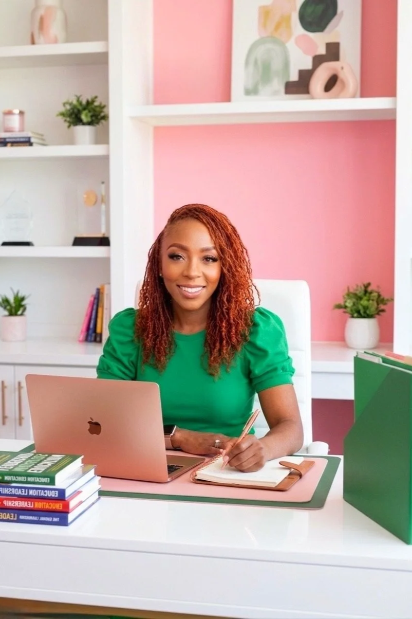 A woman with reddish-brown curly hair sitting at a desk in a colorful office or home office space. She is wearing a green top, smiling, and writing in a notebook. On the desk are a MacBook laptop, a few stacked books, a green file folder, and office supplies. The background features pink and white walls, decorative shelves with plants, books, and art pieces.
