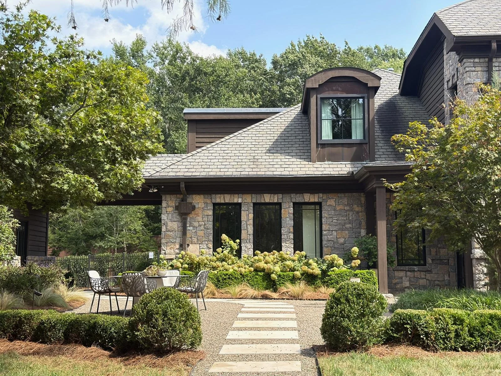 Front view of a modern house with stone and dark wood exterior, surrounded by greenery and trees, with a small garden patio in the foreground. Ultra luxury home for sale in Nashville, TN,  at $12.3M.