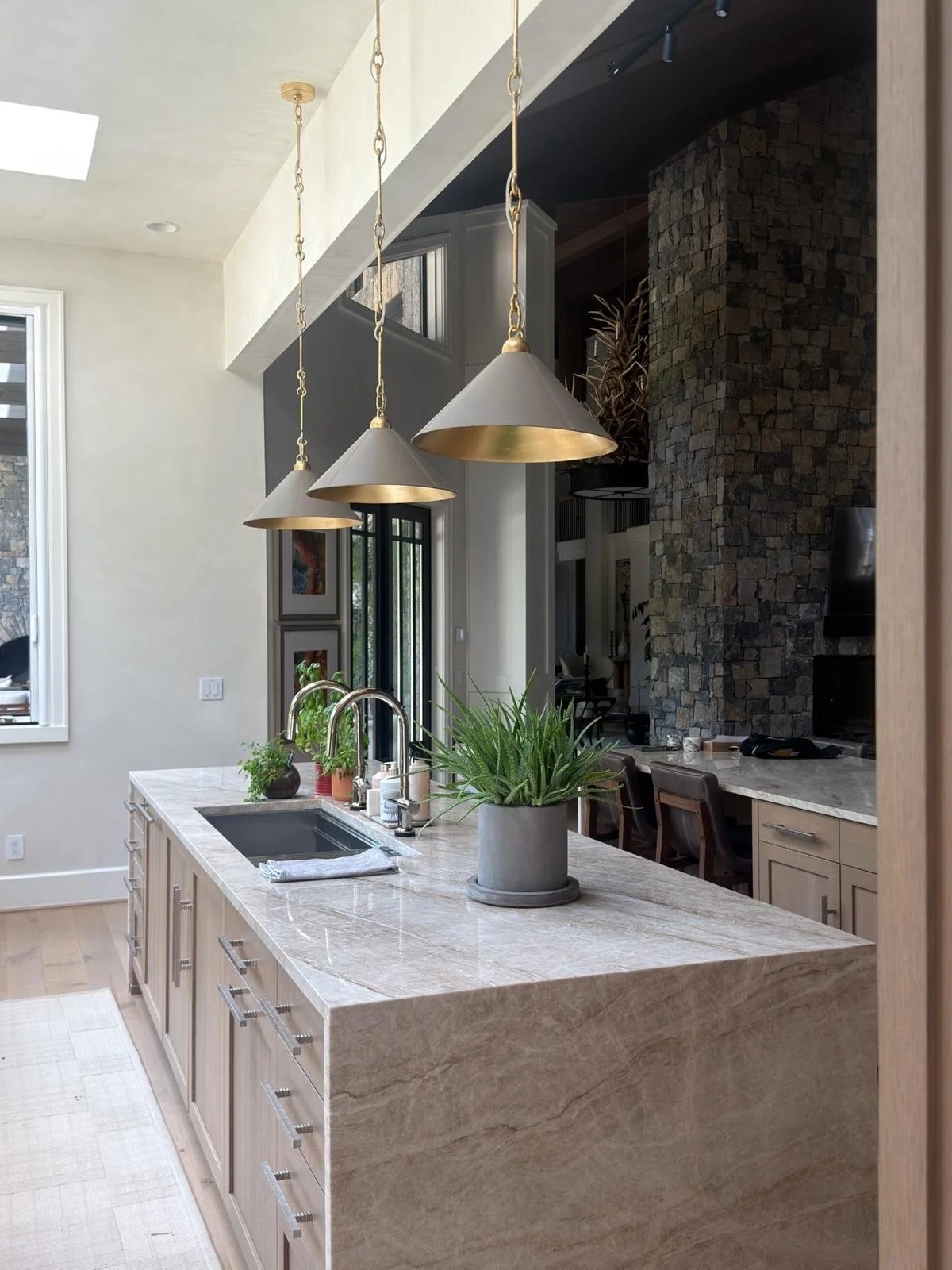 Modern kitchen with a marble island, sink, and potted plant, illuminated by three hanging pendant lights with gold accents, a stone feature wall, and large windows letting in natural light. Ultra luxury home for sale in Nashville, TN,  at $12.3M.