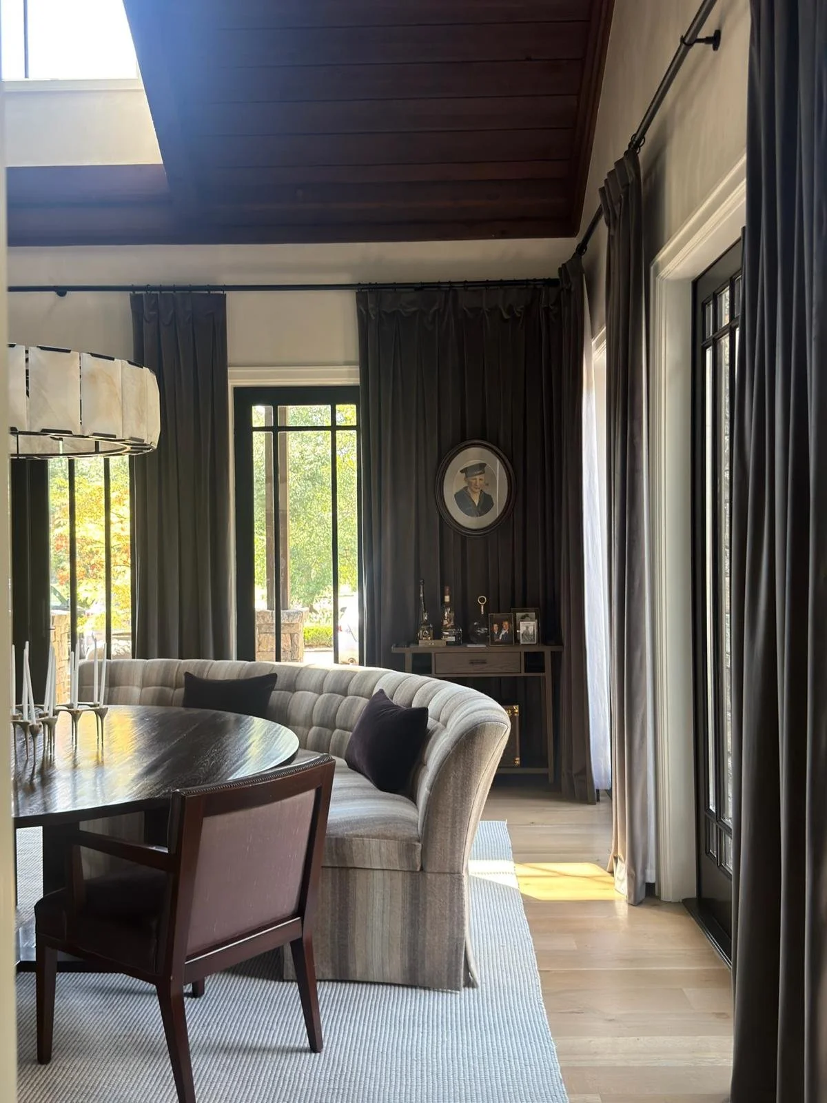 A cozy living room with large windows and dark curtains, a dining table with chairs, a curved sofa with pillows, and a small wooden sideboard with framed photos and decorative items. Ultra luxury home for sale in Nashville, TN,  at $12.3M.