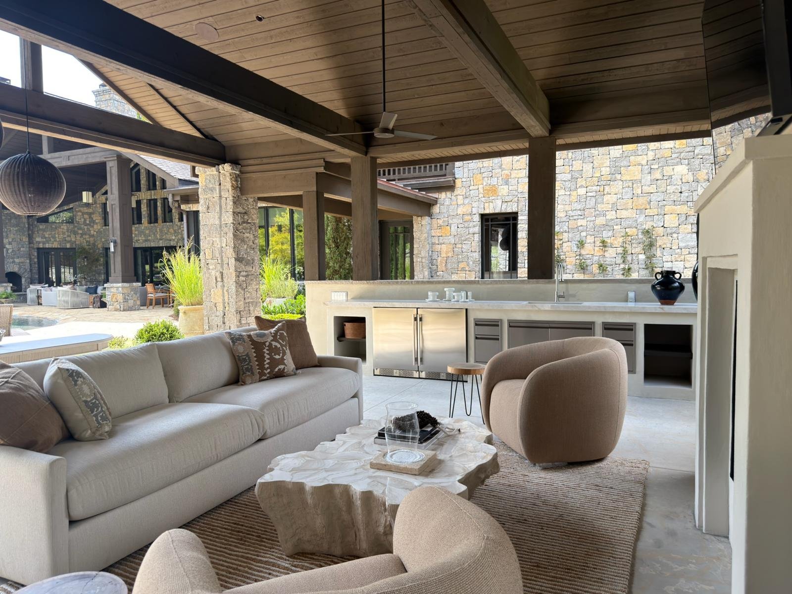 Living area with a cream sofa and beige armchair, a large stone coffee table, an overhang with a ceiling fan, and an outdoor kitchen counter with a sink, black vase, and dishes in a modern open-air patio. Ultra luxury home for sale in Nashville, TN.