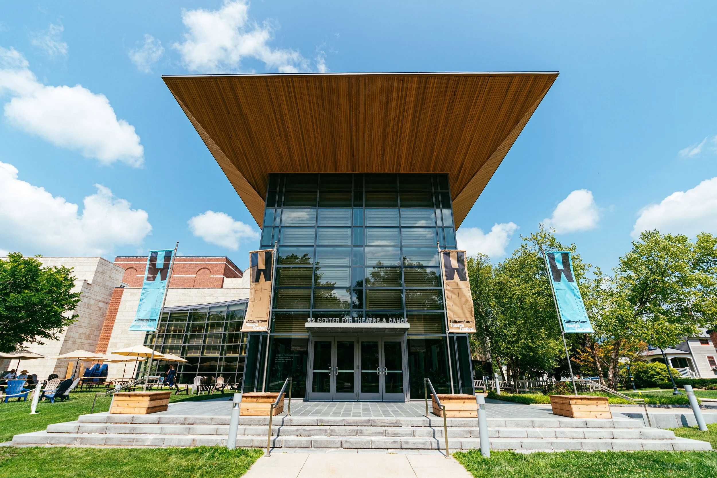 Front view of a modern theater building with a large wooden overhang, glass entrance doors, and banners reading 'Williamstown Theatre Festival' in an outdoor setting with trees and seating areas.