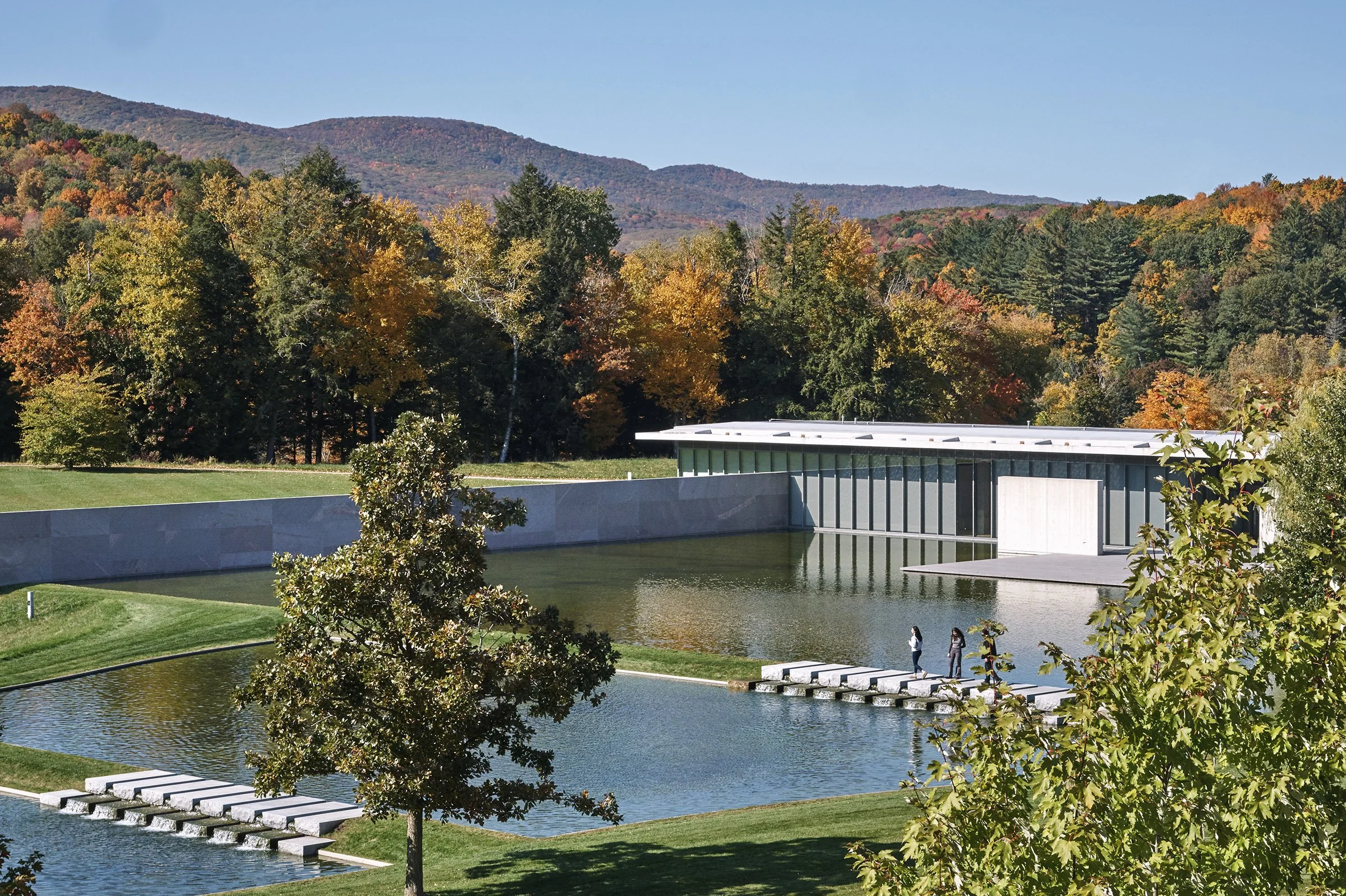 A modern building next to a pond with stepping stones, surrounded by trees with autumn foliage and mountains in the background on a clear day.