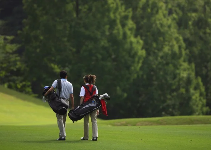 Two people walking on a golf course, carrying golf bags, with a lush green background and trees.