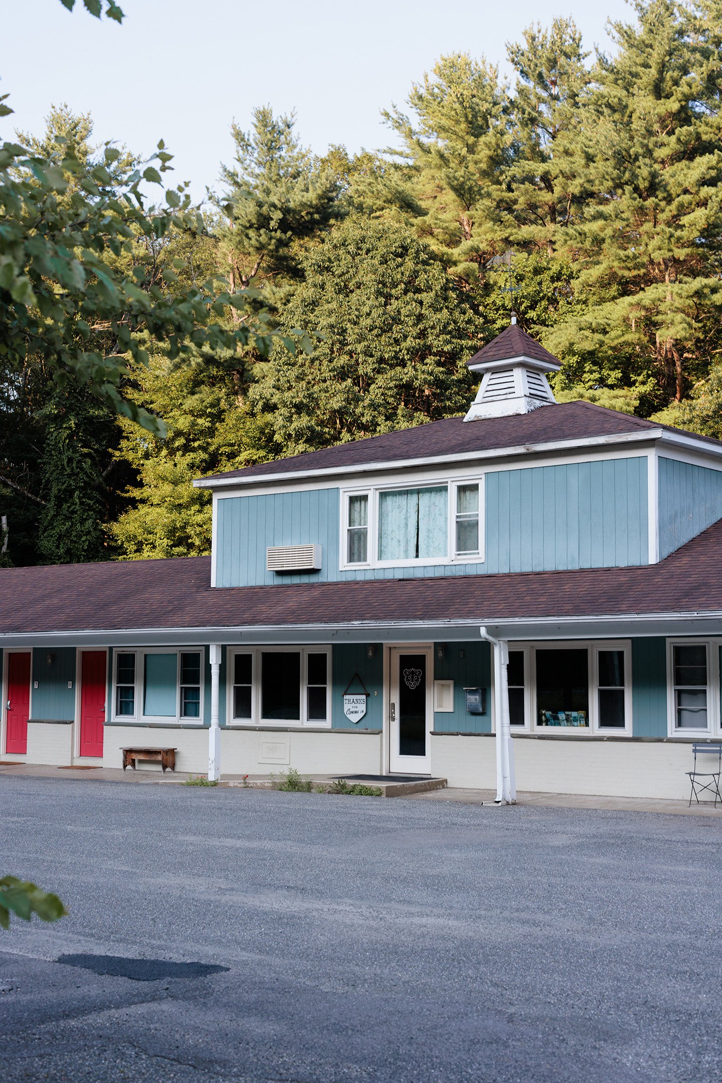 A two-story building with a light blue exterior, red doors, and a brown roof. The building has a small tower with a vent on top and an American flag on a pole. There is a parking lot in front of the building and greenery in the background.