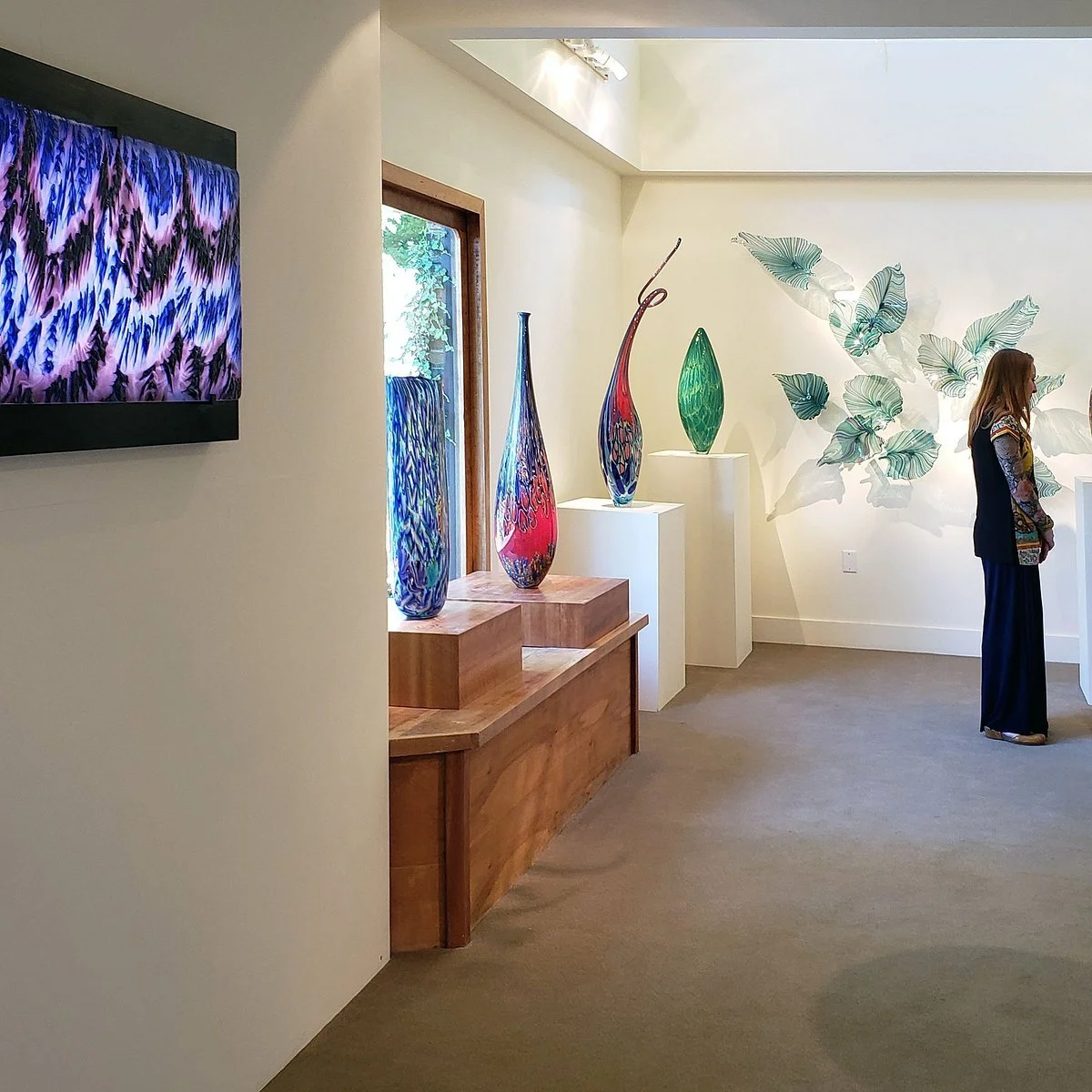 An art gallery features colorful glass vases on white pedestals, a wall decorated with large green leaf-shaped artworks, a woman in black pants and a patterned top viewing the exhibit, and a flat-screen TV displaying an abstract purple and black pattern.