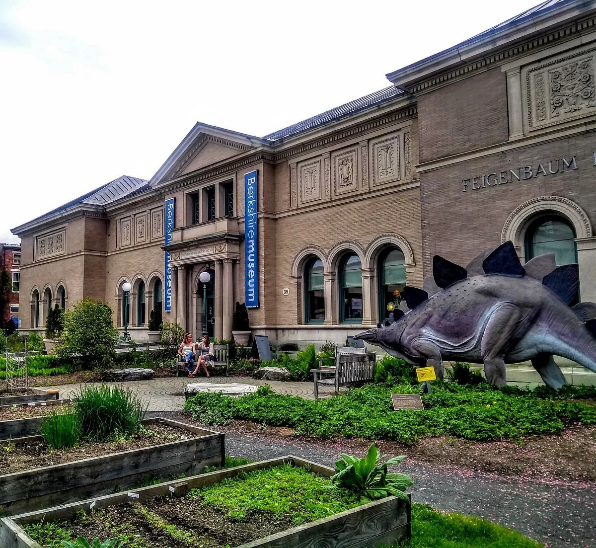 Exterior of the Berkshire Museum with a dinosaur sculpture in the garden and two women sitting on a bench.