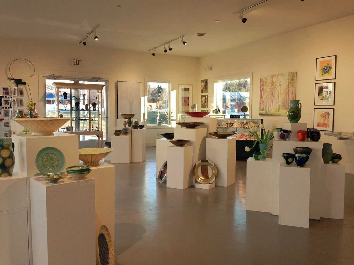 Art gallery with ceramic and glass display pieces on white pedestals, paintings on the walls, and artwork near windows.