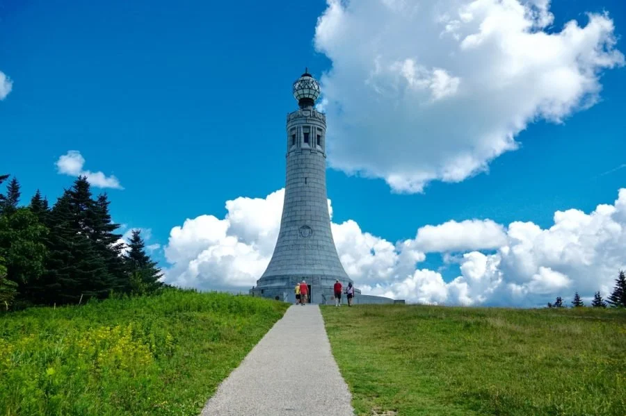 A white lighthouse standing on a grassy hill with a gravel path leading up to it, under a blue sky with scattered white clouds.