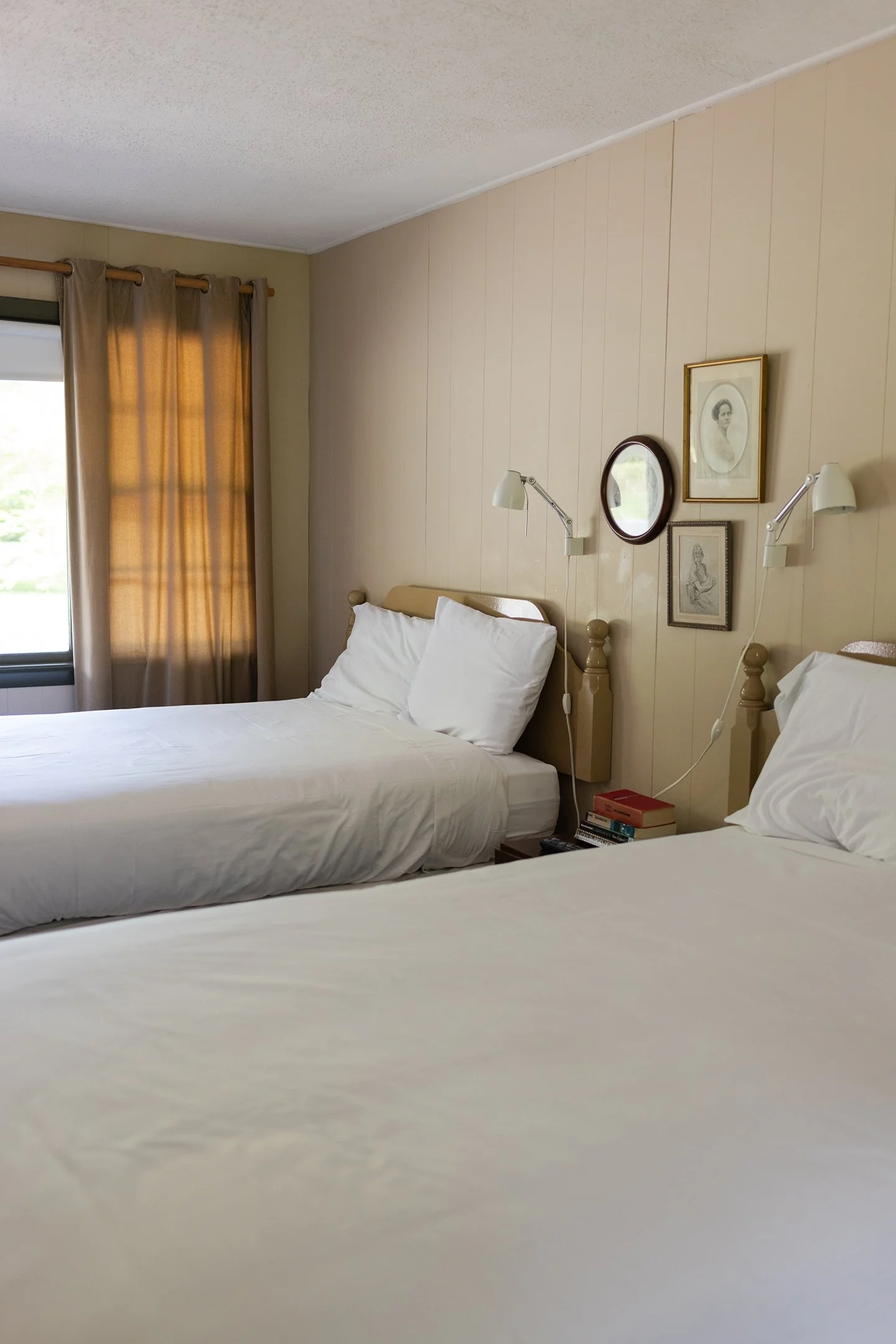 A bedroom with two beds, each with white linens and pillows. There are beige curtains on the window and picture frames on the wall above the beds. One wall has vertical wooden panels. Bedside lamps are attached to the wall.