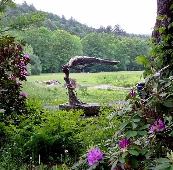 A bronze sculpture of a diver in mid-dive, positioned on a stone base in a lush garden with green foliage, purple flowers, and trees in the background.