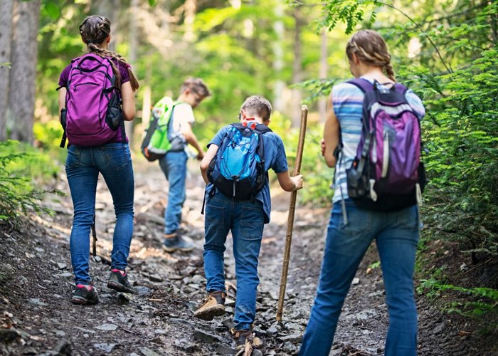 Group of children with backpacks hiking on a wooded trail, with one child holding a walking stick.