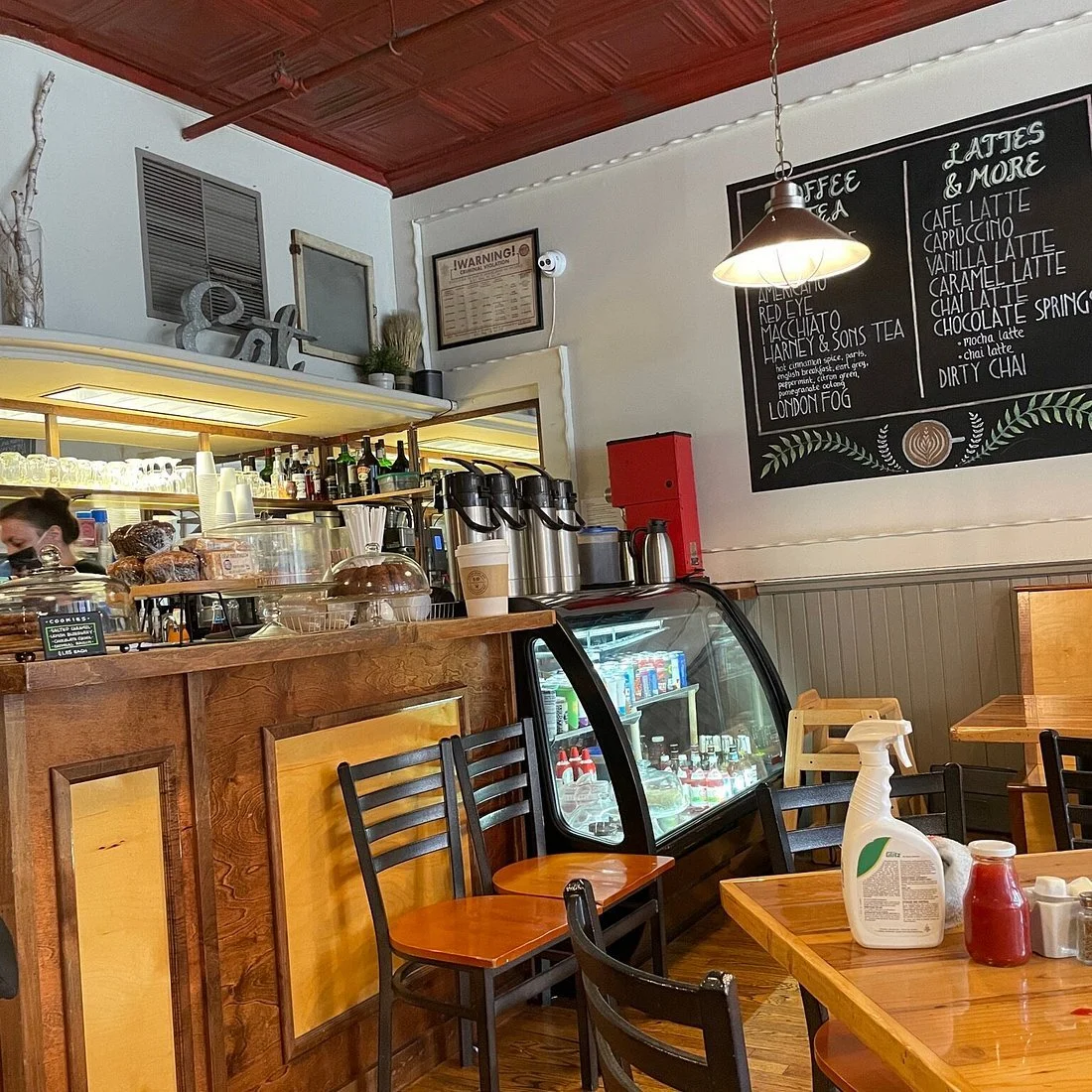Inside view of a cozy coffee shop with a wooden counter, glass display case, and chalkboard menu. Chairs and tables are arranged for customers, with condiment bottles and cleaning spray on the table.