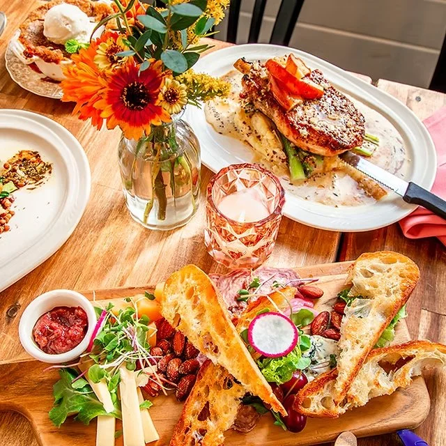 A table set with a floral centerpiece, a glass candle holder, a plate of roasted chicken with vegetables, and a charcuterie board with sliced bread, cheese, strawberries, radishes, and greens.