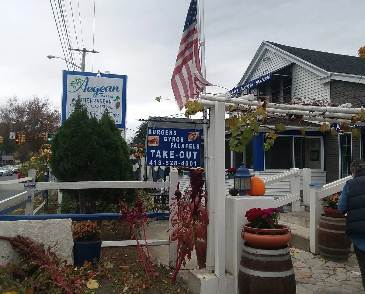 Exterior of a Mediterranean restaurant named Aegean Breeze with signs advertising burgers, gyros, falafel, and take-out, decorated with pumpkins and autumn plants, American flag flying outside.