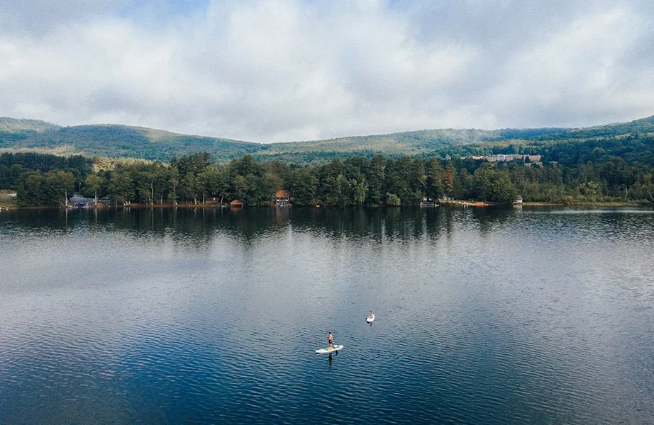 Two people paddleboarding on a calm lake surrounded by trees and rolling green hills.