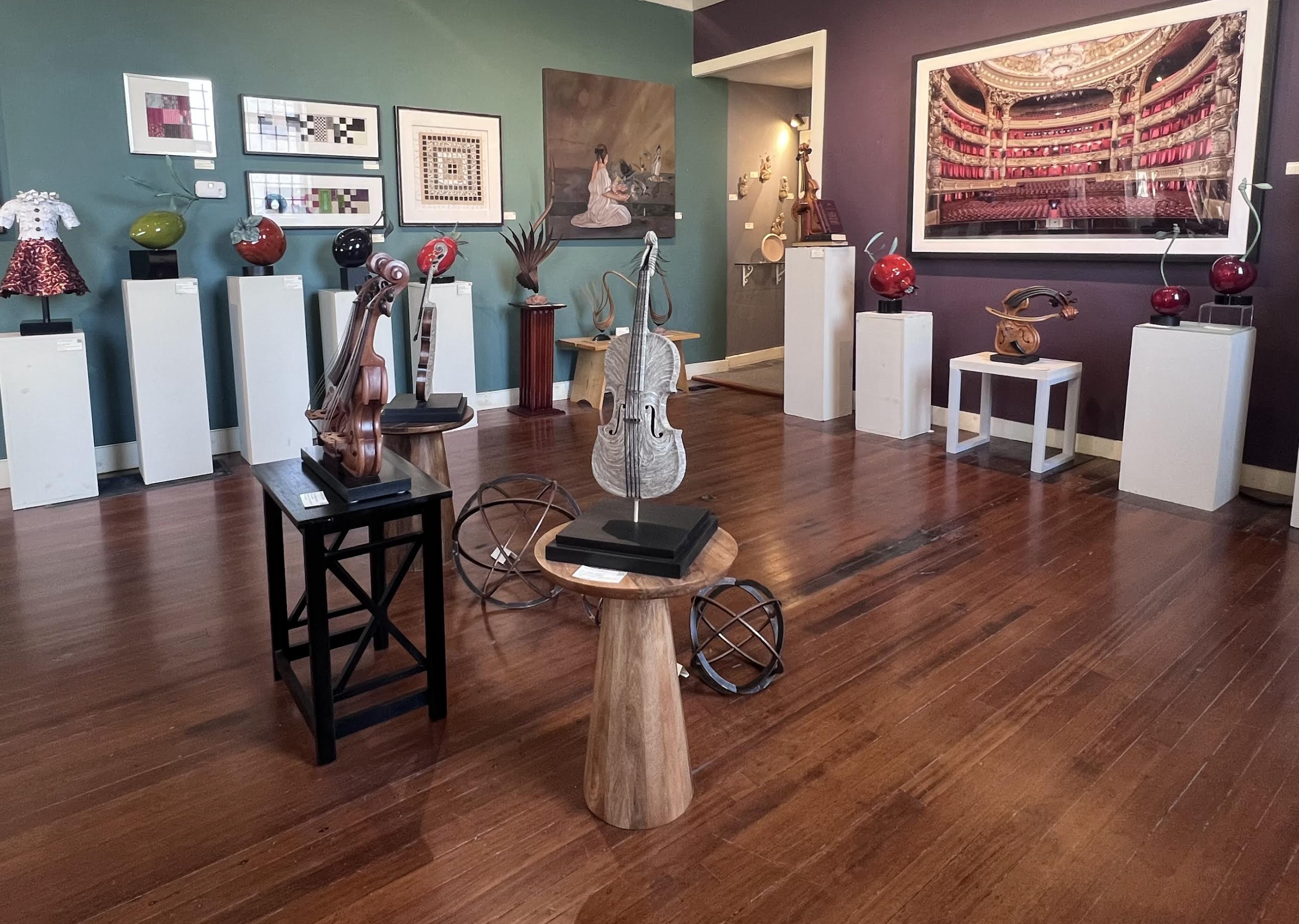 An art gallery exhibiting various artworks including paintings, sculptures, and glass art. The gallery features wooden flooring and colorful walls with a large framed picture of an opera theater or grand auditorium.
