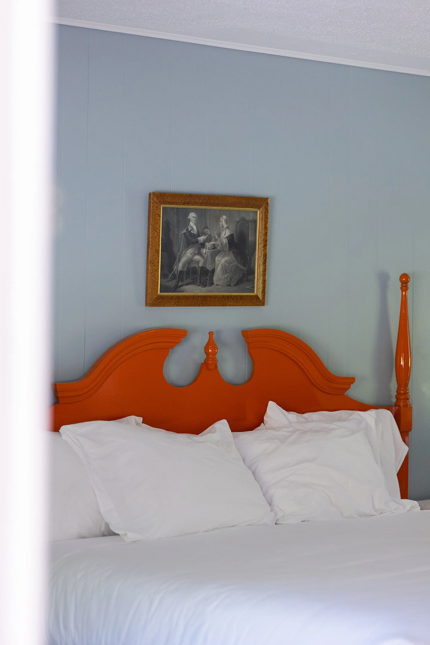 A hotel bed with white linens and pillows, a wooden headboard painted orange, and a framed painting of two people in historical clothing hanging on a light blue wall above the bed.