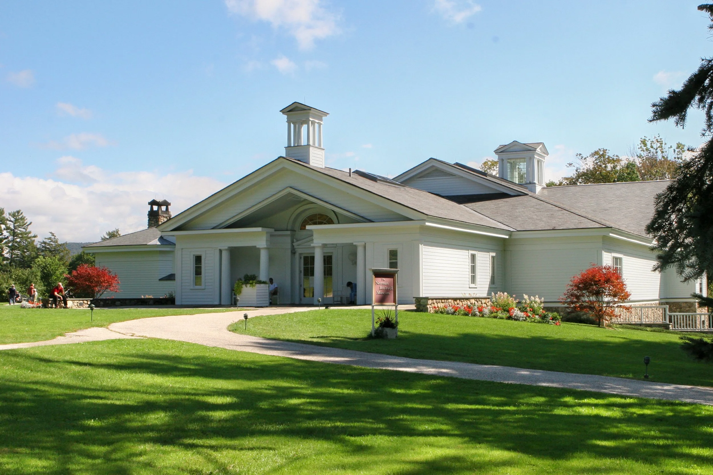 White historic building with columns and cupolas, surrounded by green lawn and trees, with people sitting and walking outside on a sunny day.