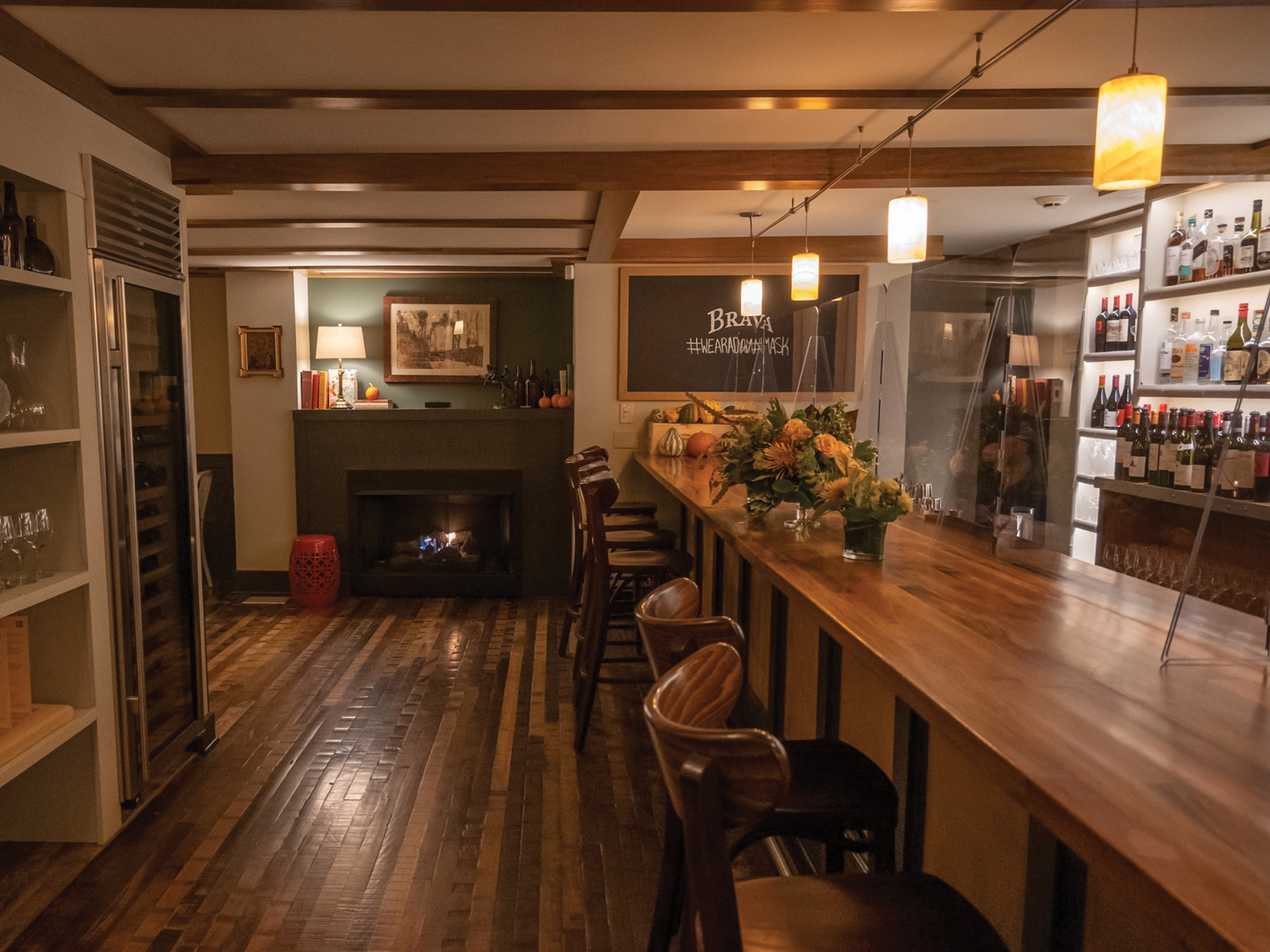 A cozy, dimly lit bar with a long wooden counter and high chairs, bottles of liquor on shelves, and a fireplace with a floral arrangement on the counter nearby.