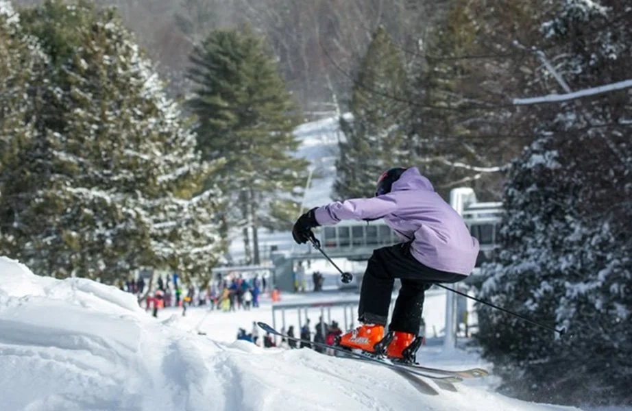 Skier descending a snowy slope in a forested area with other skiers and a ski lift in the background.