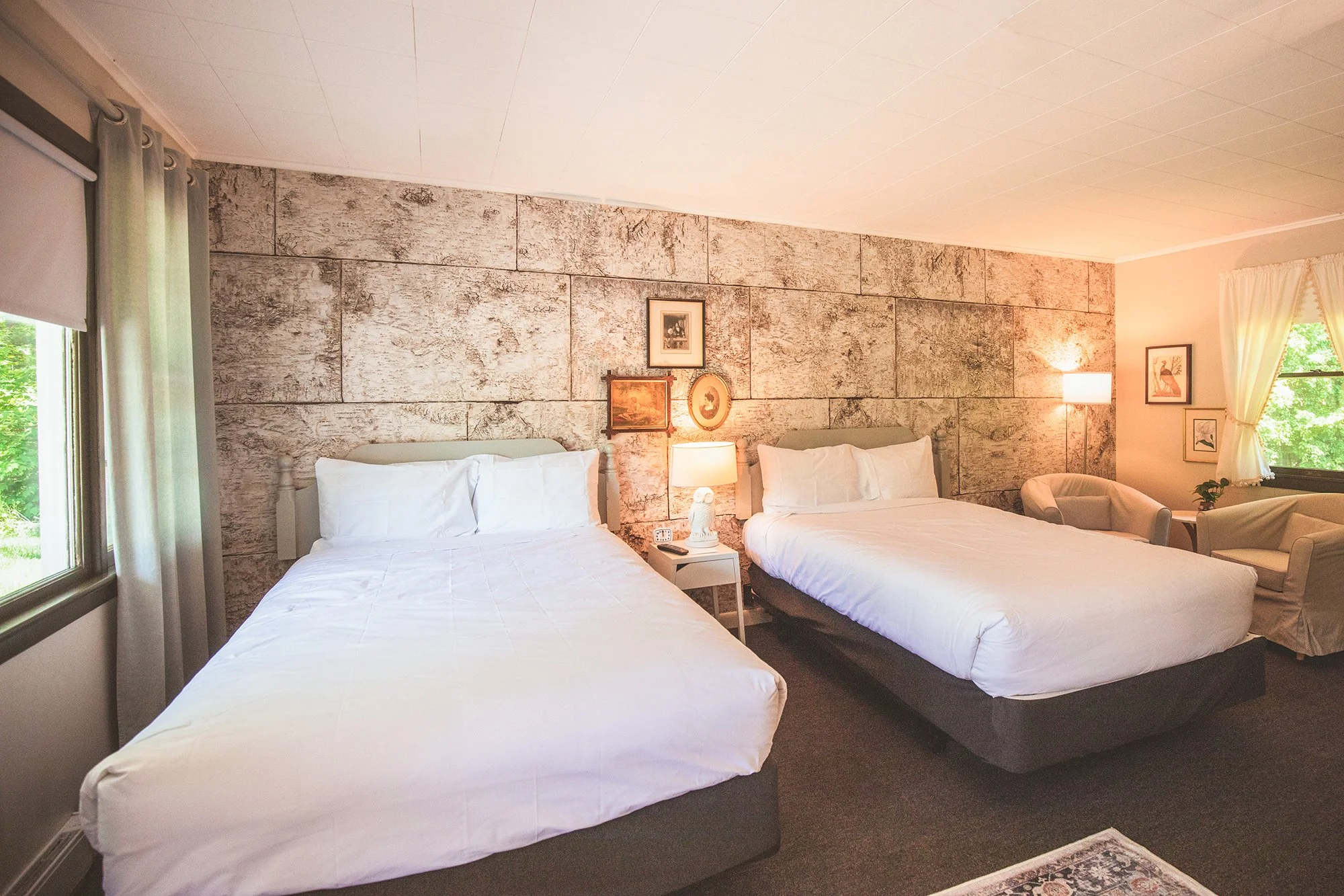 Two beds in a warmly lit hotel room with a stone accent wall, framed artwork, and windows with curtains, one showing greenery outside.