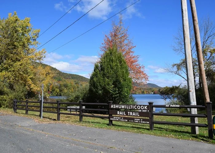 A scenic view of Ashwulitcoock Rail Trail with a wooden fence, trees in fall foliage, and a lake in the background on a clear day.