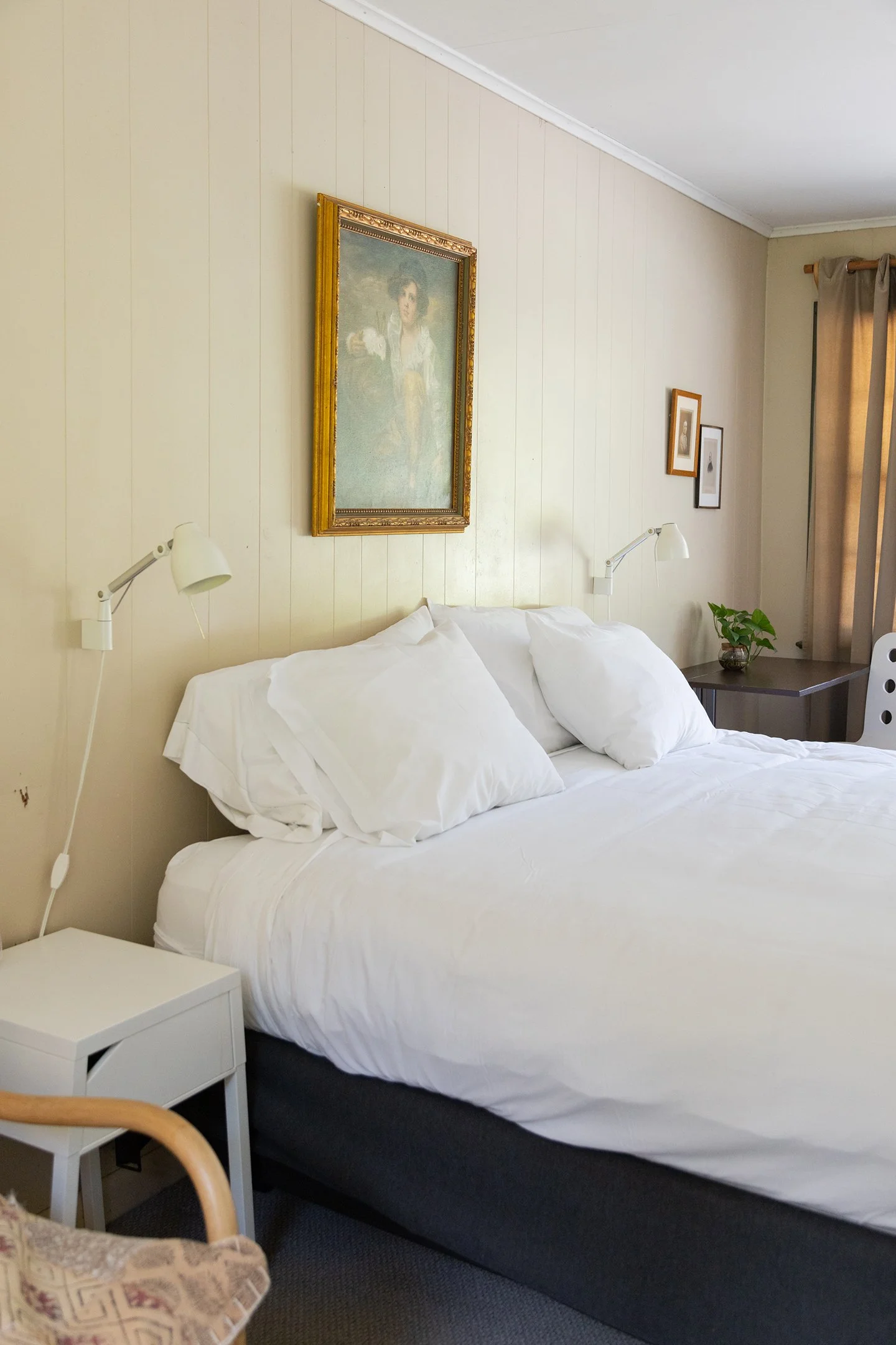 A cozy bedroom with a bed featuring white linens and pillows, a nightstand with a plant, and artistic wall decor including a large framed portrait, small framed pictures, and a window with curtains.